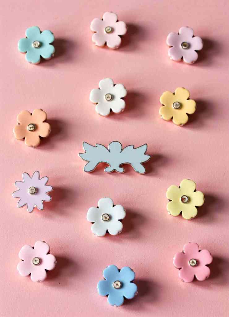 A delightful arrangement of small flower-shaped enamel pins in soft pastel hues.

The pins, including blue, pink, white, and yellow, are set against a pink background. A decorative blue-winged enamel pin is centrally placed, adding a whimsical touch.