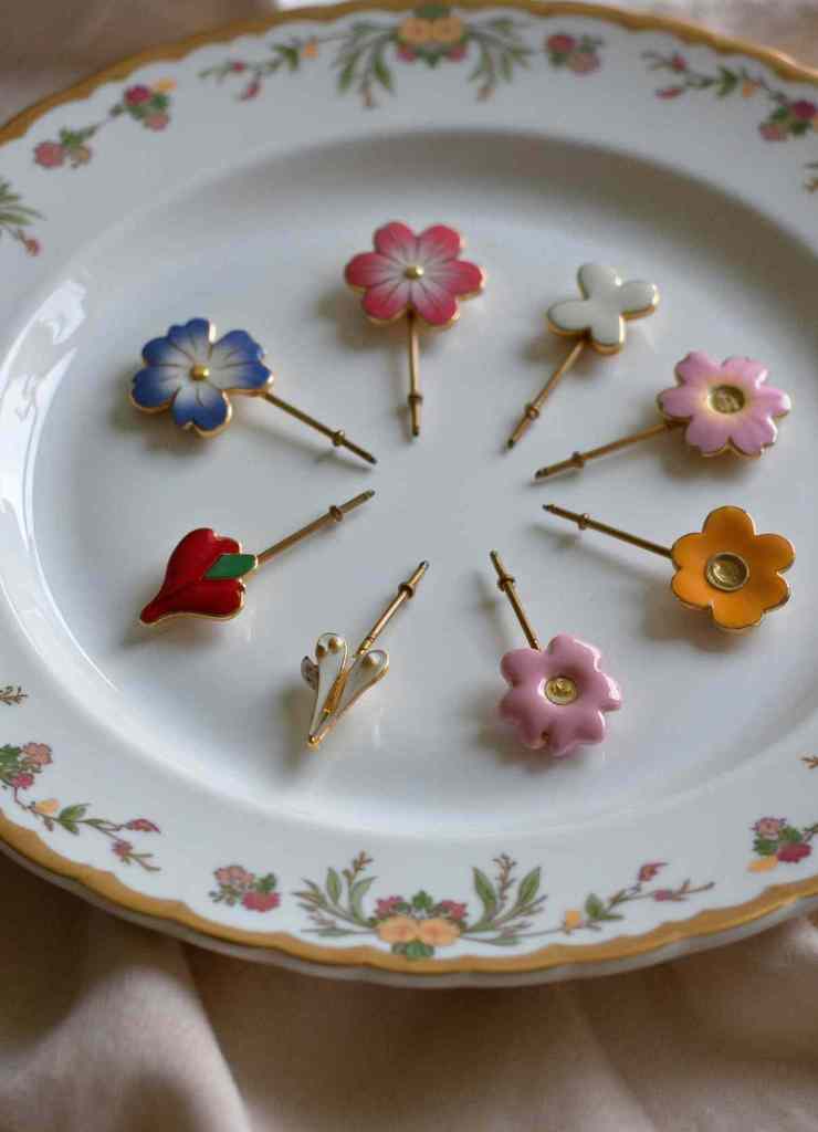 A collection of delicate floral stick pins arranged in a circular pattern on a vintage-style floral plate. Each pin showcases unique colors, including blue, pink, yellow, and red, with gold details, making them perfect for adding a refined touch to any outfit.