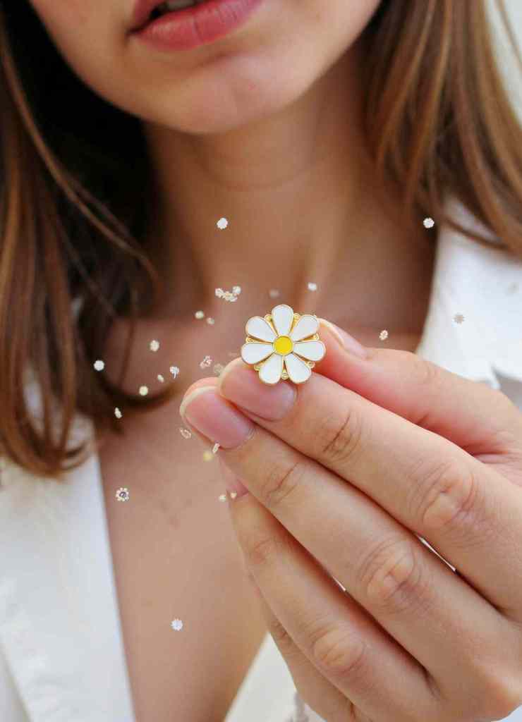 A close-up of a woman holding a white daisy enamel pin with a yellow center. Tiny floating flowers add a magical effect, emphasizing the charm of this classic, minimalist piece.