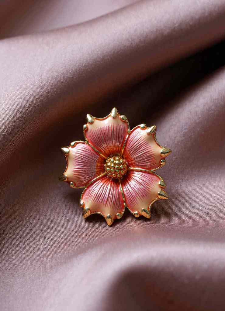 A beautifully crafted pink enamel flower brooch with gold-tipped petals is showcased against a luxurious satin background. The soft, warm tones create an elegant and timeless appeal.