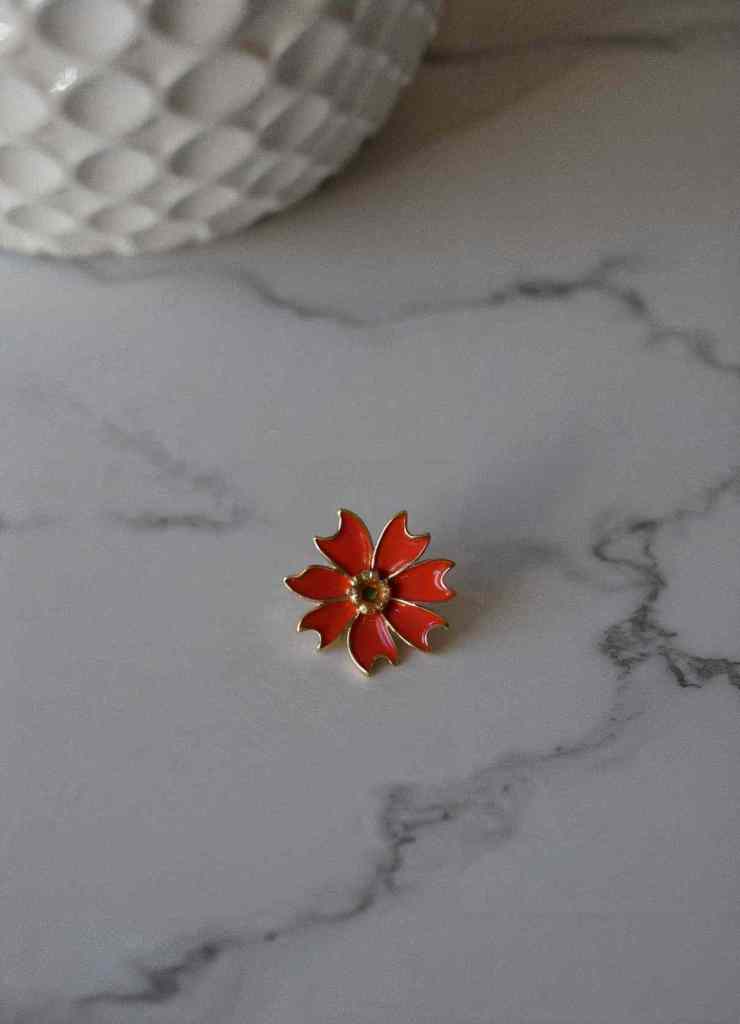 A vibrant red enamel flower pin with gold detailing sits on a marble surface. Its sleek and bold design makes it a versatile accessory for both modern and vintage-inspired outfits.