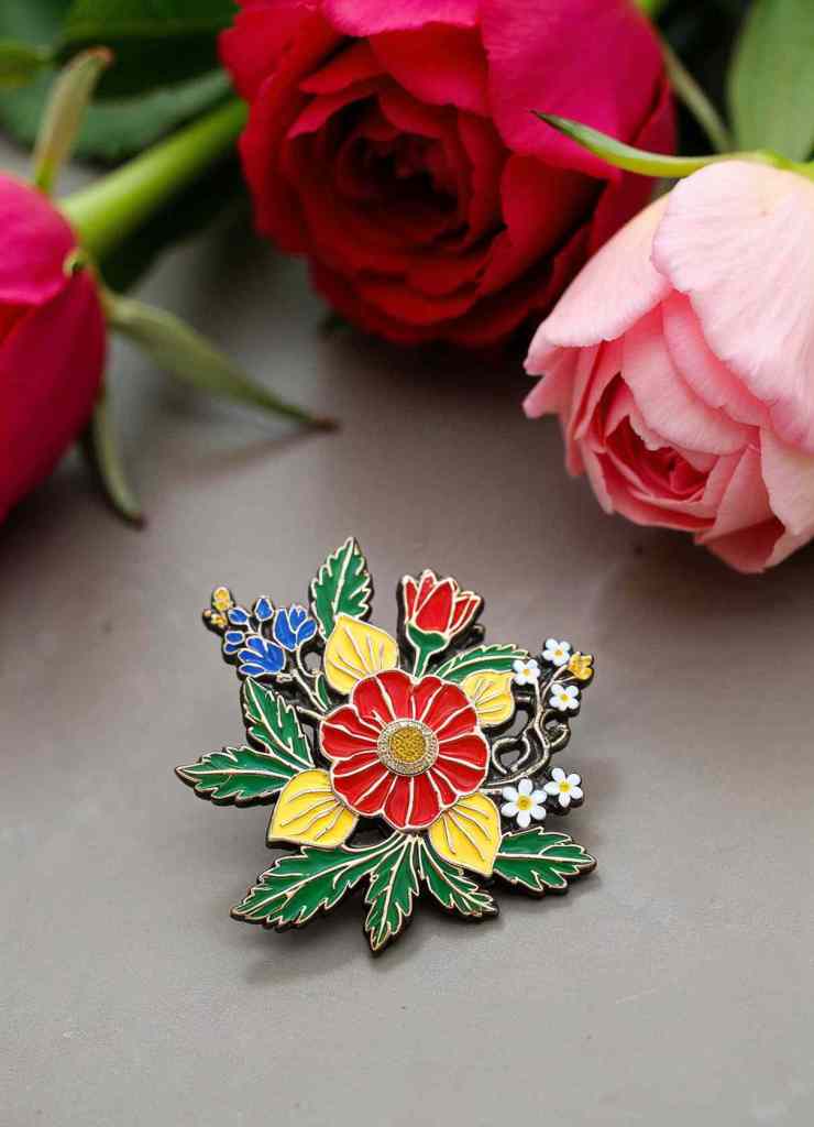 A bouquet-inspired enamel brooch featuring red, yellow, blue, and white flowers with intricate green leaves. The brooch is placed alongside real roses, emphasizing its botanical elegance.