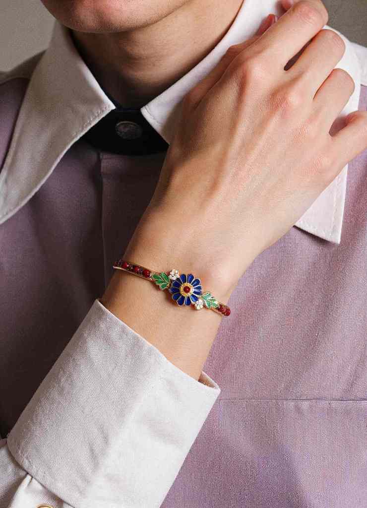 A close-up of a dainty bracelet featuring a blue enamel flower with red and green accents. The intricate detailing adds a vintage charm, making it a timeless piece for any jewelry collection.