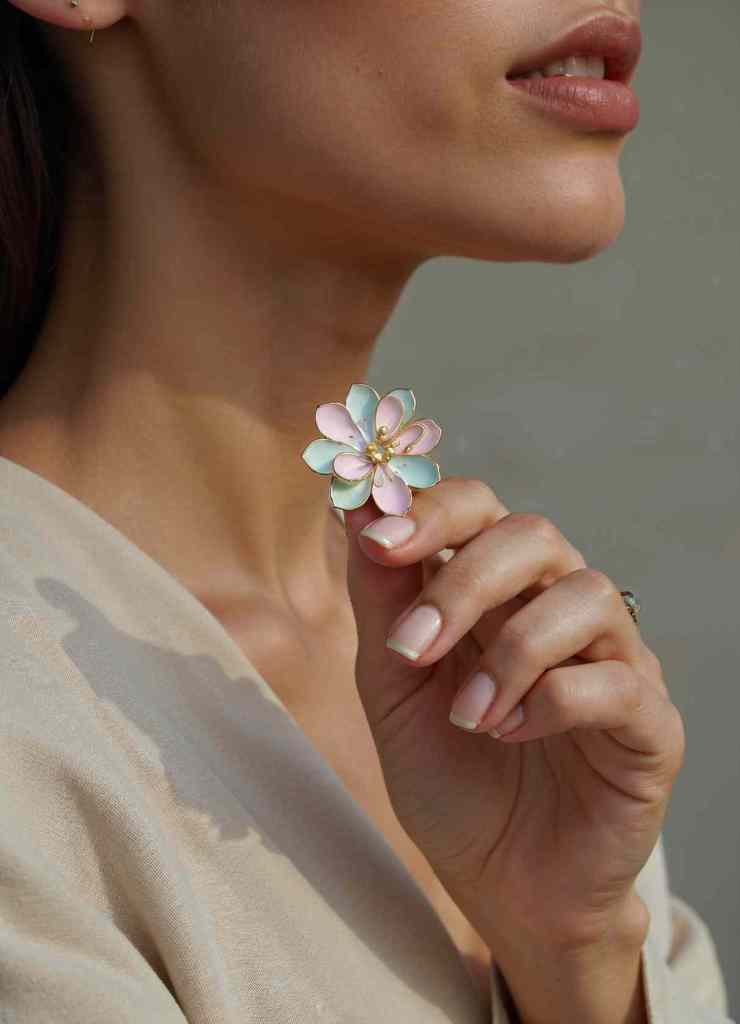 A close-up of a woman delicately holding a pastel-colored enamel flower ring pin.

The soft pink and blue petals are outlined in gold, with golden accents at the center, exuding a sophisticated yet charming look.