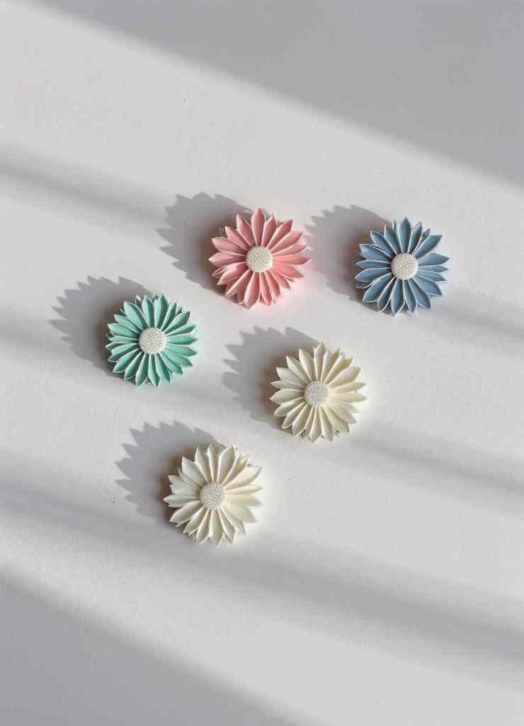 A collection of pastel daisy enamel pins in pink, blue, green, and white shades. The pins are arranged in soft natural lighting, highlighting their delicate textures and intricate petal details.