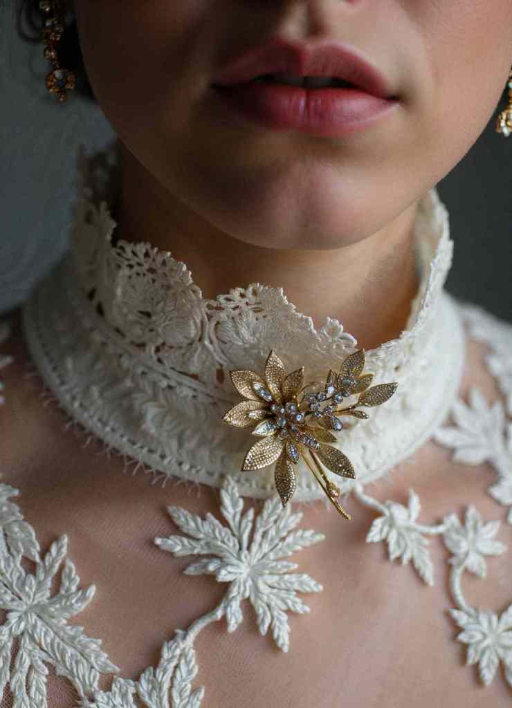 A stunning golden floral brooch adorned with shimmering rhinestones sits elegantly on the high lace collar of a white embroidered dress.

The brooch’s intricate filigree leaves and floral patterns enhance its vintage appeal.