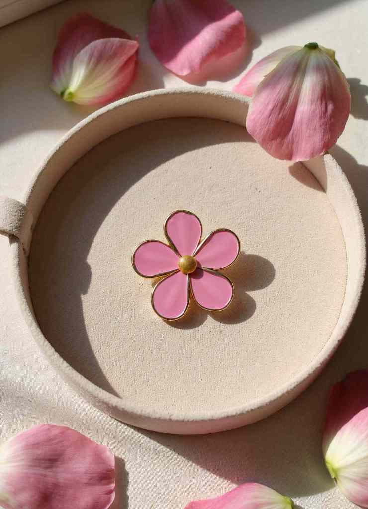 A beautiful pink flower enamel pin with gold edges is displayed in a round velvet jewelry case, surrounded by delicate pink rose petals.

The soft lighting highlights the pin’s glossy finish and romantic aesthetic.