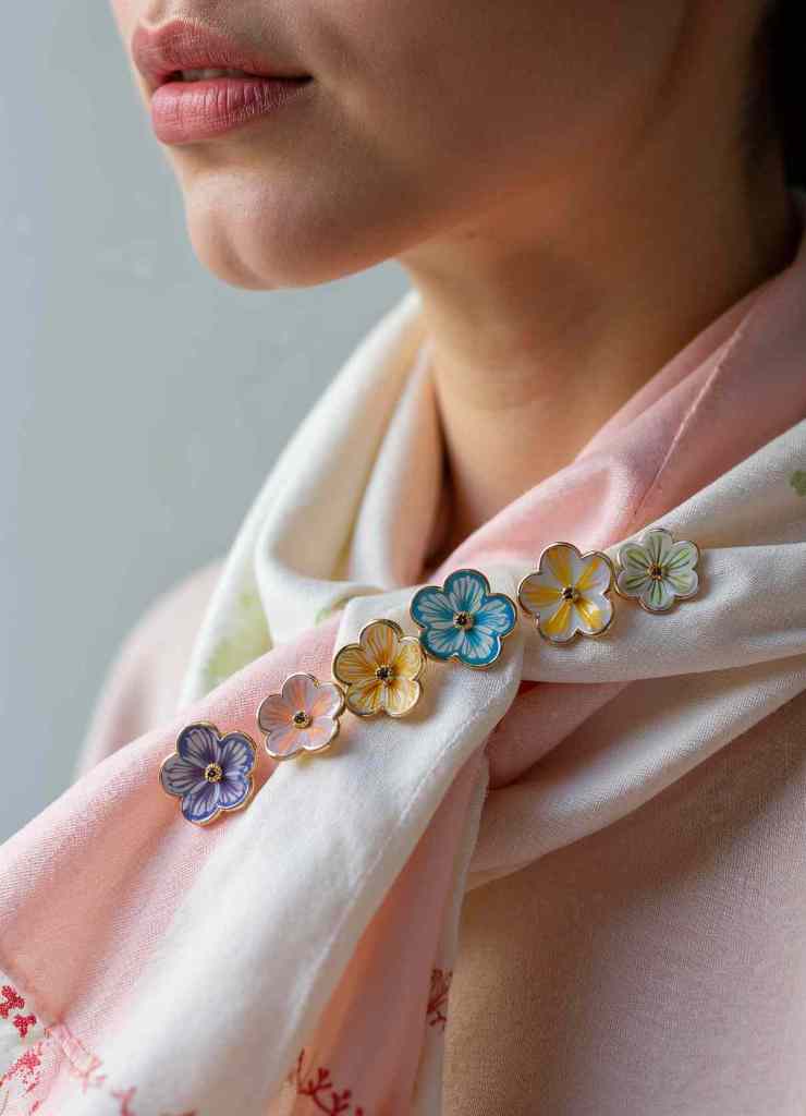 A woman drapes a pastel silk scarf adorned with multicolored enamel flower pins.

The floral pins, in hues of blue, yellow, pink, and purple, enhance the scarf’s elegance, creating a stunning accessory arrangement.