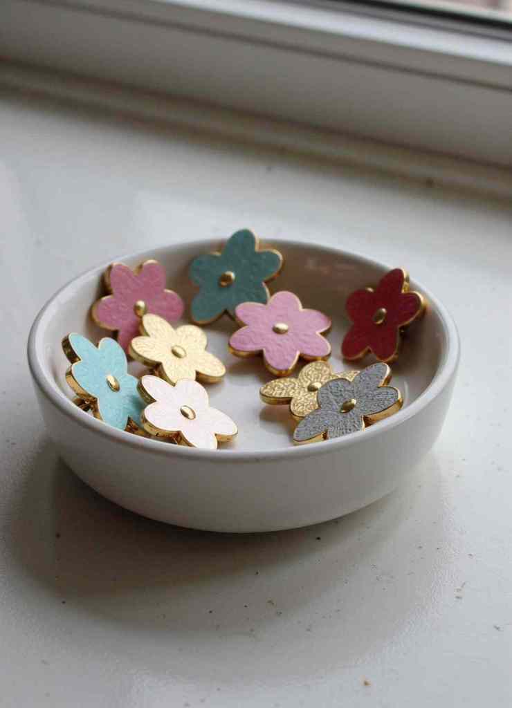 A collection of pastel-colored flower enamel pins with gold edges is displayed inside a simple white ceramic bowl near a window.

The combination of matte and glossy textures adds depth to these charming accessories.