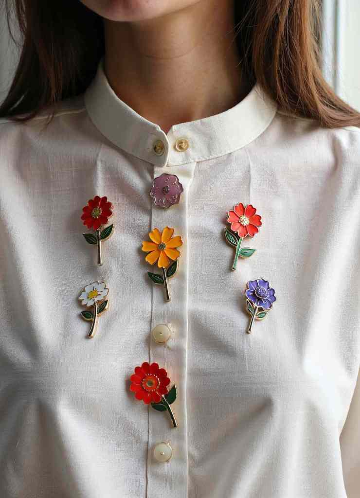 A collection of vibrant enamel flower brooches with green stems is pinned onto a white button-up shirt.

The colorful flowers, ranging from red to orange, yellow, and purple, create a cheerful and playful aesthetic.