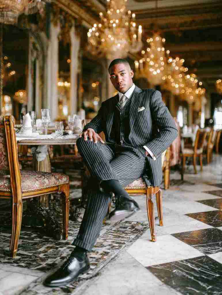 A well-dressed man in a pinstriped suit sits confidently at a table in an elegant restaurant with chandeliers and a luxurious ambiance.