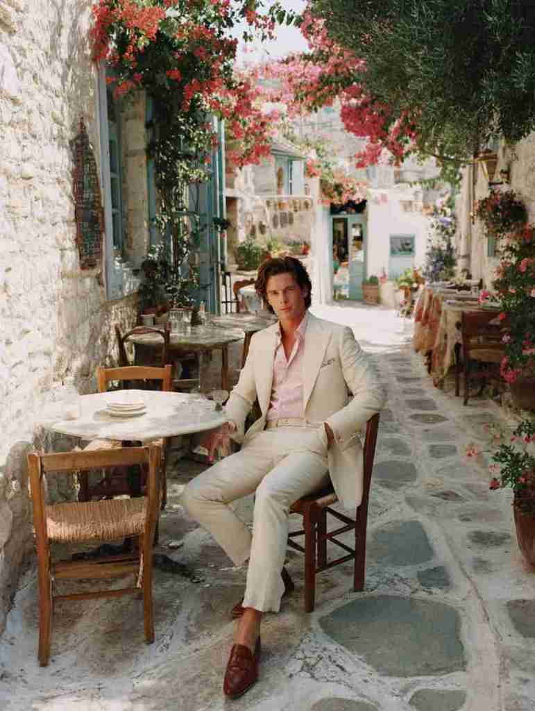 A well-dressed man in a light suit and pink shirt sitting at a small outdoor table adorned with flowers in a charming restaurant setting.