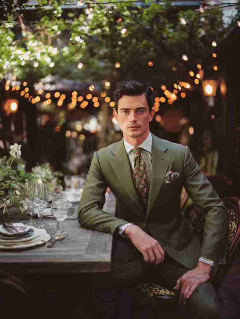 A well-dressed man in a green suit sits at a beautifully set table in an outdoor dining space, surrounded by greenery and softly lit with string lights.