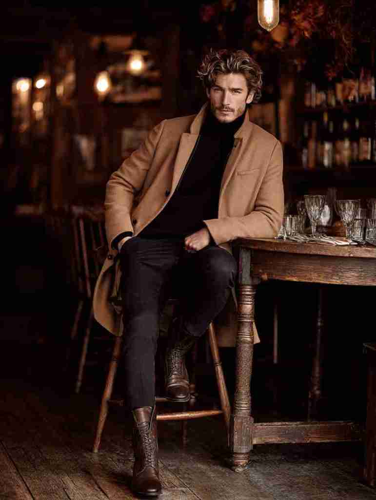 A stylish man seated at a rustic wooden table in a dimly lit restaurant, wearing a beige coat and a black turtleneck, exuding confidence and elegance.