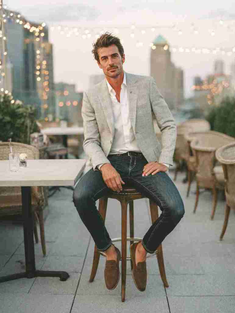 A man sitting on a bar stool at a restaurant rooftop, wearing a light gray blazer over a white shirt and dark jeans, with brown loafers, enjoying the evening ambiance.