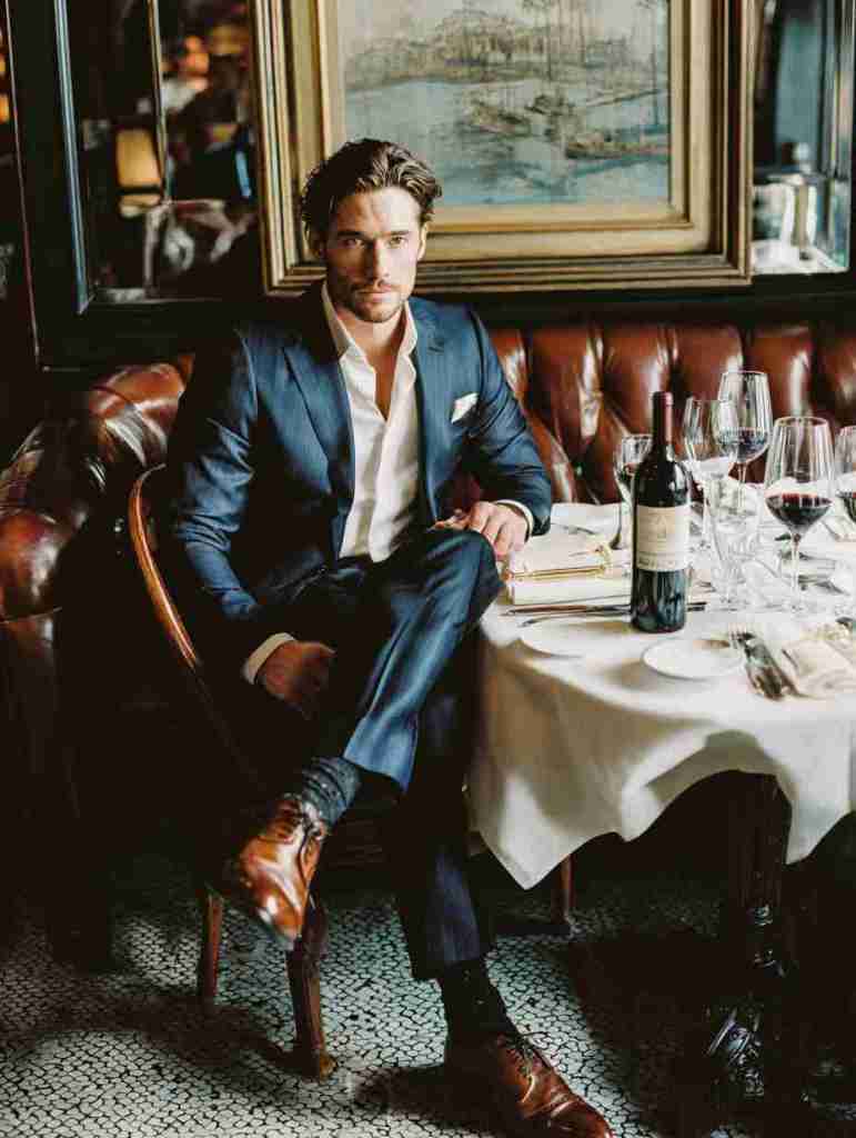 A well-dressed man seated at a table in a refined restaurant, looking confidently at the camera. The setting includes a stylish interior with a wine bottle and elegant diningware.