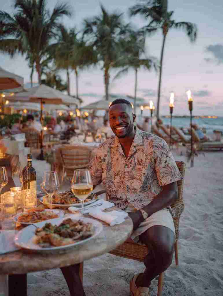 A man smiling while dining outdoors at a beach restaurant, enjoying a variety of dishes and a glass of white wine, with palm trees and a sunset in the background.