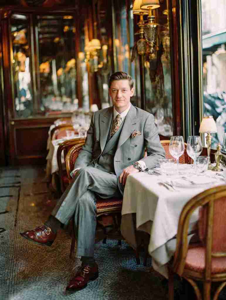 A well-dressed man sitting in an elegant restaurant, wearing a grey suit and stylish shoes, with tables set for dining in the background.