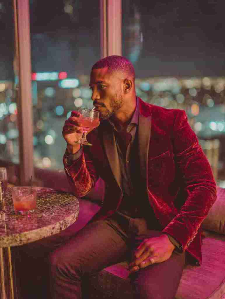 A man in a stylish red velvet blazer enjoys a drink while sitting at a bar with a city skyline view.